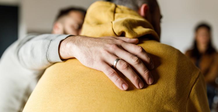 Compassionate man embracing his friend who is sharing a heavy story regarding his mental health during a group therapy session.
