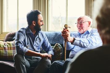 Young male doctor and elderly man sitting on sofa and smiling during care home visit